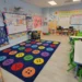 Bright preschool classroom at The Little Butterflies with a colorful circle-time rug, child-size tables, and STEM, math, and language wall displays